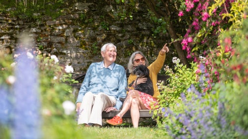 Two people are sitting on a wooden bench in a lush garden filled with various colorful flowers and greenery. One person is wearing a light blue sweater and beige pants, while the other is wearing an orange cardigan, red floral skirt, and orange shoes. The second person is holding a small black dog on their lap and pointing towards something in the distance. The background features a stone wall covered with moss and plants.
